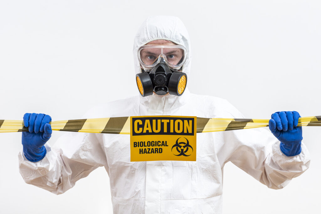 Man in hazmat suit and mask standing behind a sign that says "caution - biological hazard"